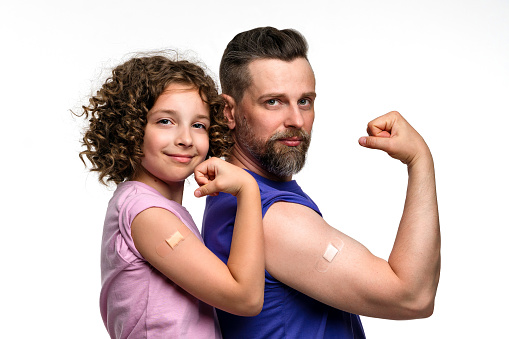 Family in basic t-shirts showing biceps with band-aid after vaccine injection. Dad and daughter 10-12s wear pink and violet t-shirts on white studio wall. Antiviral immunization. Revaccination