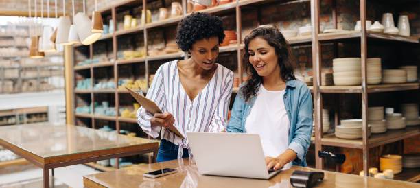 deux jeunes commerçants utilisant un ordinateur portable dans leur magasin - retail photos et images de collection