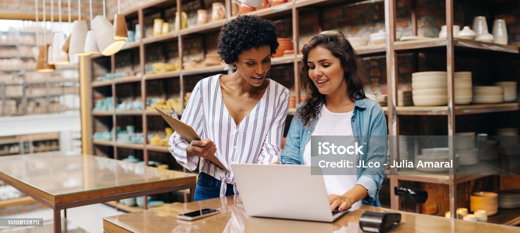 Two young shop owners using a laptop in their store Two young shop owners using a laptop while working in a ceramic store. Female entrepreneurs managing online orders on their website. Young businesswomen running a creative small business together. Small Business Stock Photo