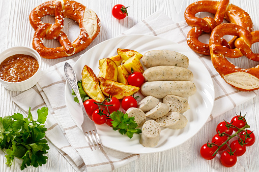 Weisswurst, bavarian white sausage of minced veal, pork back bacon, spices and parsley on white plate with roast potato, fresh tomatoes, soft pretzels, sweet mustard, horizontal view from above
