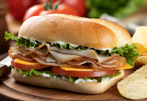 Submarine sandwich made with turkey, cheese, lettuce, tomato and mayonnaise with potato chips on wooden platter