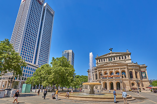 Frankfurt am Main, Germany - July 08, 2022: Pedestrians on the Opernplatz in front of the Old Opera in Frankfurt. In the foreground the Lucae Fountain. In the background a skyscraper from the financial district.