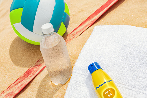 A multi-colored beach volleyball sitting in the sand next to the red court boundary tape, a bottle of cold water and a bottle of spray on sunscreen on a white towel.
