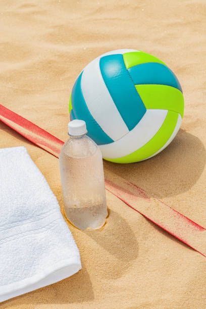 A beach volleyball sitting next to a towel and a bottle of water in the sand stock photo