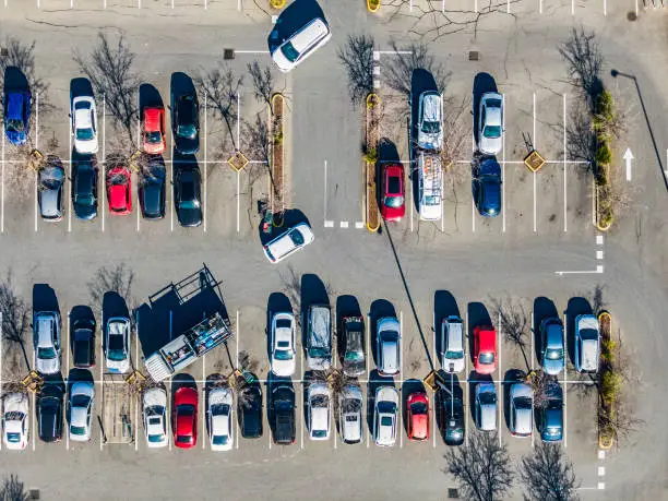 Aerial view directly above suburban shopping centre carpark; trade truck parked diagonally across 3 spaces; cars arriving and exiting parking spaces, mostly red, blue, white and grey cars, direction arrow, bare trees, shadows.