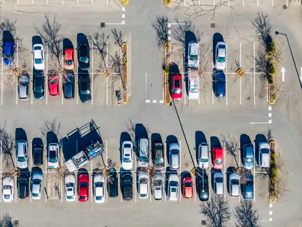 Aerial view directly above suburban shopping centre carpark; trade truck parked diagonally across 3 spaces; all vehicles stationary in parking spaces, mostly red, blue, white and grey cars, direction arrow, bare trees, shadows.