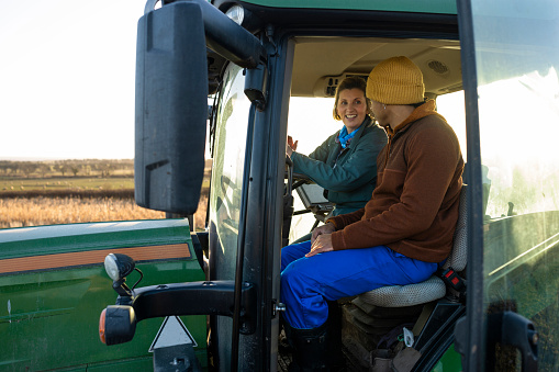 A mid length side view of a female farmer sitting with her young male apprentice farmer sitting in the cab of the tractor. She is teaching him about the controls on the tractor and how to use it.