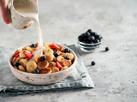 Trendy food - pancake cereal. Milk pouring on heap of mini cereal pancakes in bowl. Tiny cereal pancakes with berries in craft plate over gray cement background. Copy space for text or design.