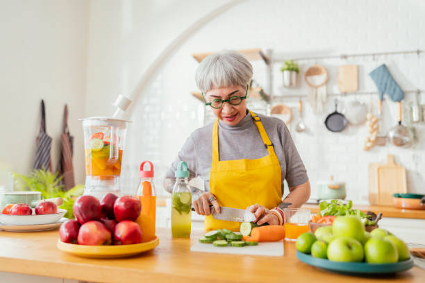 reife lächelnde tattoo-frau, die salat, obst und gemüse isst. attraktive reife frau mit frischem grünem obstsalat zu hause. senior frau schürze steht in der küchentheke entspannt im haus - fruit salad fruit healthy eating making stock-fotos und bilder