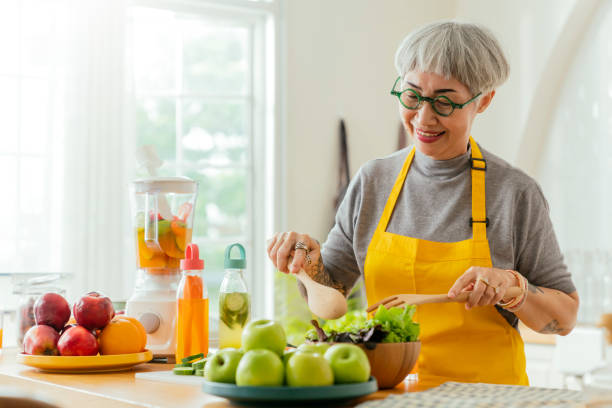 reife lächelnde tattoo-frau, die salat, obst und gemüse isst. attraktive reife frau mit frischem grünem obstsalat zu hause. senior frau schürze steht in der küchentheke entspannt im haus - fruit salad fruit healthy eating making stock-fotos und bilder