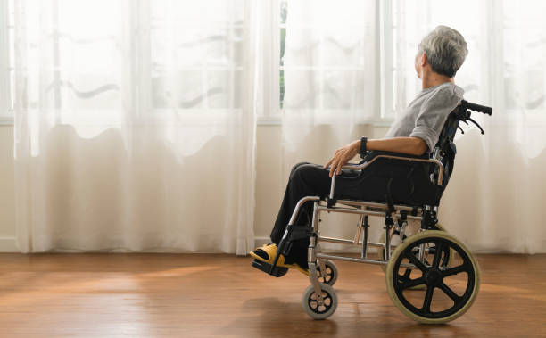 Shot of a senior man looking out of a window while sitting in a wheelchair at a home,elder asian male stay home alone in wheelchair look at view out of window from living room stock photo