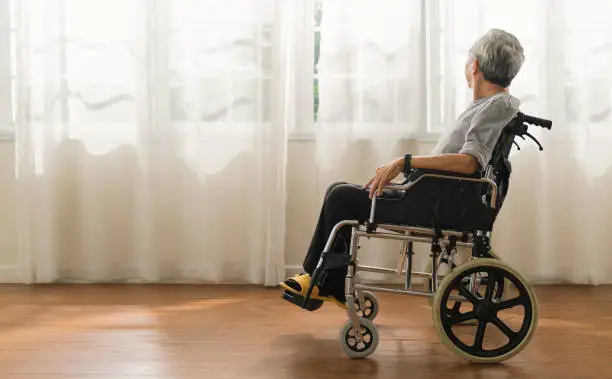 Photo of Shot of a senior man looking out of a window while sitting in a wheelchair at a home,elder asian male stay home alone in wheelchair look at view out of window from living room