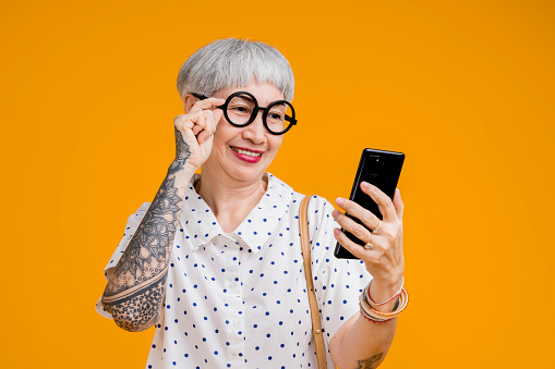 Portrait of attractive funky asian tattoo cheerful grey-haired elder old senior woman touching spectacles and smartphone device internet conected isolated over yellow pastel background color
