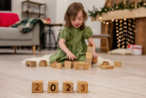 Wooden cubes with the numbers 2023 stand on the floor in a row. Background of a blurred happy girl in a green dress, who is playing, building a tower from a construction set. Child development at home
