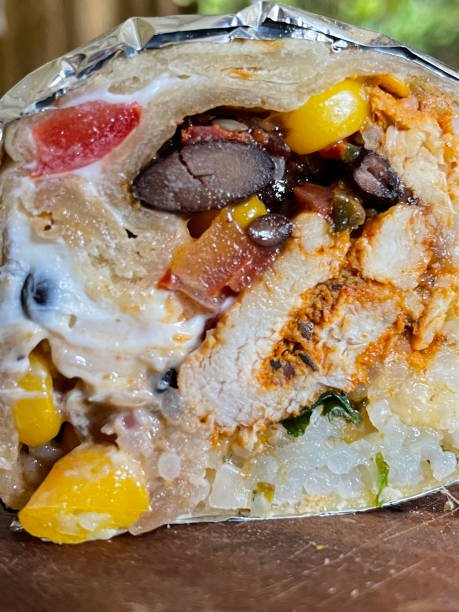 Full frame cross section image of rolled, homemade, soft wheat tortilla flatbread burrito wrapped in tinfoil, rice, raisin, marinated chicken, sweetcorn, chopped cucumber, red onion and tomato filling, on wooden chopping board, focus on foreground
