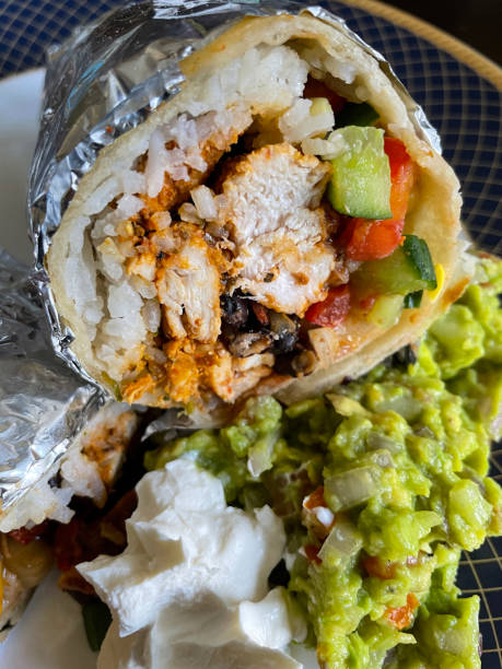 istock Full frame cross section image of rolled, homemade, soft wheat tortilla flatbread burrito wrapped tinfoil, rice, raisin, marinated chicken, sweetcorn, chopped cucumber, red onion and tomato filling, blue rimmed plate, guacamole, sour cream 1410845573