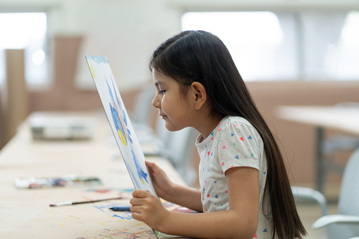 A little latin girl holding and looking at her painting very close.