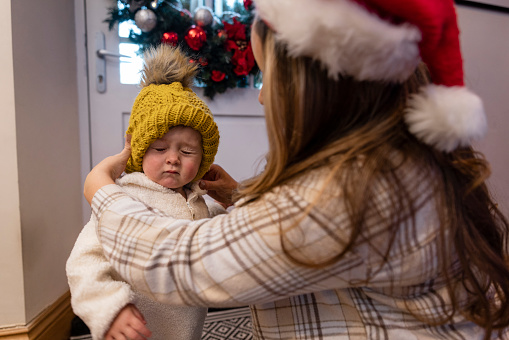 A medium shot of a woman and her toddler daughter in the entrance to their home at Christmas time. The mother is kneeling down and helping her daughter put on a knit hat before they go outside.