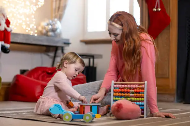 A medium shot of a woman and her toddler daughter wearing casual clothing in their living room at Christmas time. They are sitting on the floor as they play with educational toys.
