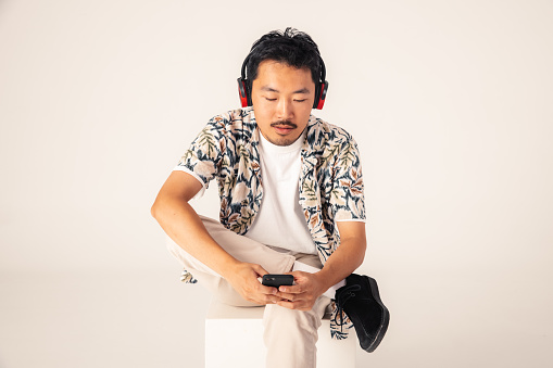 East Asian model going for portraits photos in studio. In this photo, he is sitting and enjoy listening content using headset and his phone.