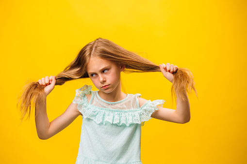 Artistic girl shows her emotions. The child stands on a yellow background, in a beautiful turquoise dress. Kid holds hair with hands. child and copy space. yellow background