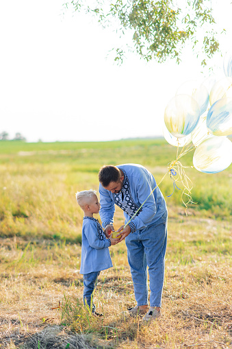 Happy father with his son plays on meadow with air balloons. They are dressed in Ukrainian national embroidered shirts.