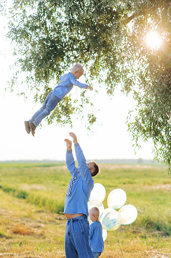 Happy father plays with his son and throws his up during walk on the meadow, while other his son plays with air balloons near him. They are dressed in Ukrainian national embroidered shirts.