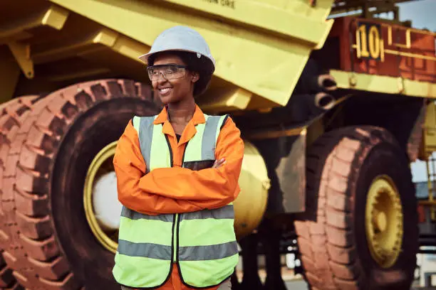 A young African woman mine worker is standing in front of a large haul dump truck wearing her personal protective wear