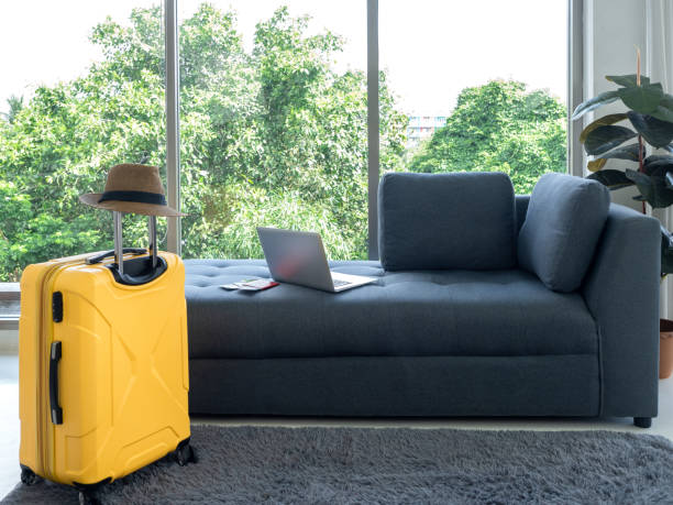 istock Empty grey cozy fabric sofa seat with laptop computer and yellow suitcase with beach hat near huge glass window and green plant pot decoration in living room. Ready to summer vacation travel concept. 1410861197