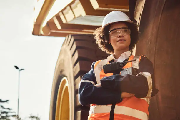 A young African woman mine worker is standing in front of a large haul dump truck wearing her personal protective wear