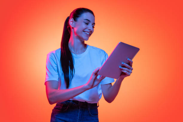 retrato de jovem emocional, estudante de camiseta branca isolada em fundo de cor laranja em luz de neon. conceito de beleza, moda, emoções - electrical equipment technology elegance computer - fotografias e filmes do acervo