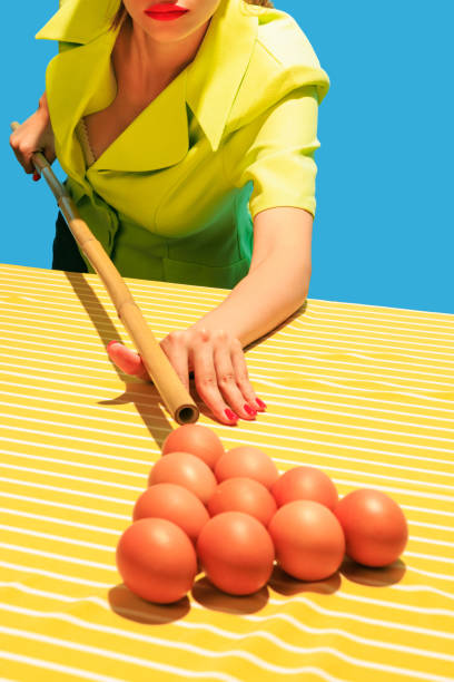 Creative image of female hands playing billiards, hitting egg balls with cue isolated on blue yellow background