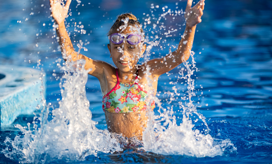 A girl in bright pink swimsuit and black swimming goggles, funny jumping into deep blue pool with clean transparent cool water against background of the warm summer sunny sunset