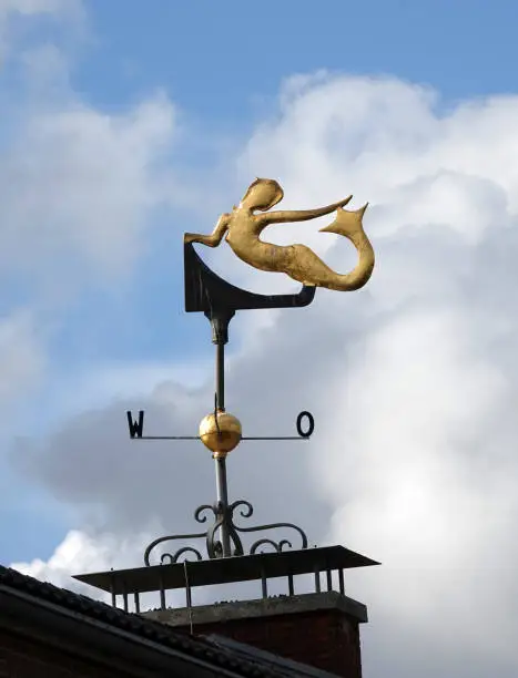 Gold colored weather vane in the shape of a mermaid. Cloudy sky in the background