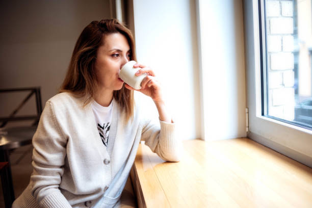 une jeune femme séduisante dans un café boit du thé ou du café et regarde par la fenêtre - restaurant business person setting the table clothing photos et images de collection