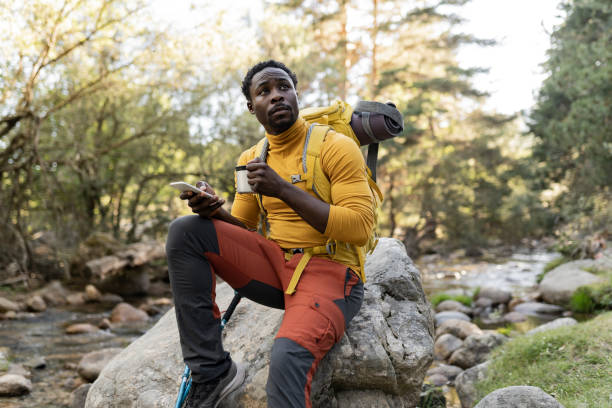 istock handsome young african american man with backpack smiling at sunset sitting on the rock, while holding a mug with drink. 1410870787
