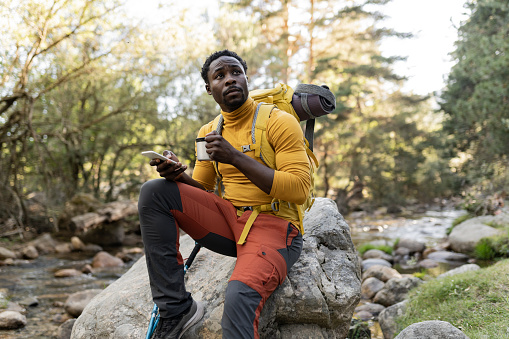 handsome young african american man with backpack smiling at sunset sitting on the rock, while holding a mug with drink.