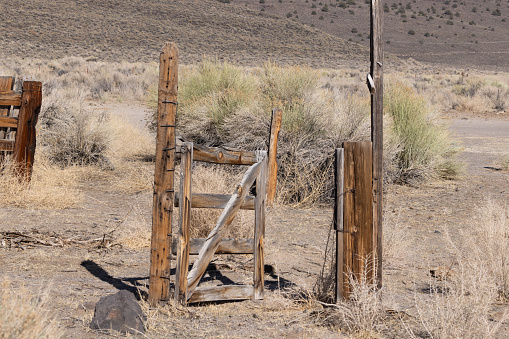 Dried sagebrush and sand surround old wood fence post with tattered open gate. Old rusty barbed wire is cut and hanging. The dry and desolate desert of the Sierra mountain range of Mono Country, California