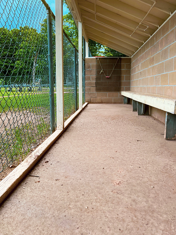 Empty little league baseball or softball dugout. Long view of players bench and chain link fence overlooking the dirt field.