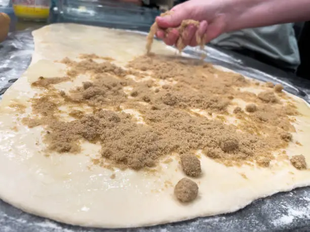 Preparing dough with butter and brown sugar for rolling up and baking into cinnamon rolls. Hand is gently spreading brown sugar onto dough