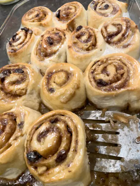 Four rows of hot sticky cinnamon rolls with nuts and raisins and shinny glaze. Metal spatula removing one fresh cinnamon roll from glass pan