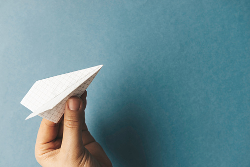 Hand is holding white paper plane in front of blue background.