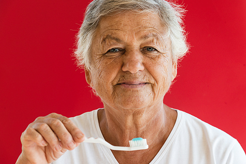 Caucasian senior woman is holding  white toothbrush with blue toothpaste in front of his face in front of red background with a warm smile. Representing dental health for senior adults concept