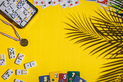 White dominoes with colorful dots in a metal box, winner medal, playing cards, ligretto, uno lie in a round frame on a yellow background with a shadow of a palm tree branch and copy space in the center, flat lay close-up. Concept summer board game.