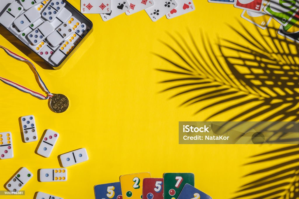 White dominoes, playing cards, uno, ligretto and winner medal on yellow background. Board game. White dominoes with colorful dots in a metal box, winner medal, playing cards, ligretto, uno lie in a round frame on a yellow background with a shadow of a palm tree branch and copy space in the center, flat lay close-up. Concept summer board game. UNO City Complex Stock Photo
