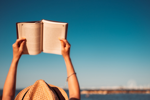 Woman holding a book in the air, reading on the beach.