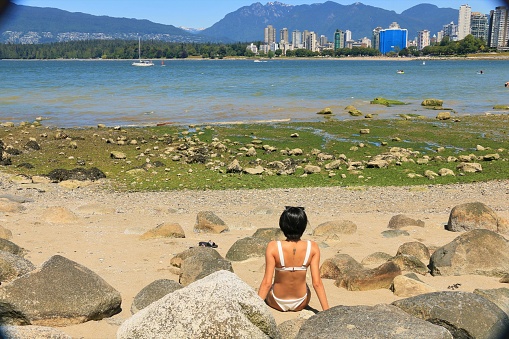 A Mexican woman sitting no a beach looking onto English Bay and Vancouver, BC, Canada with mountains in the background.