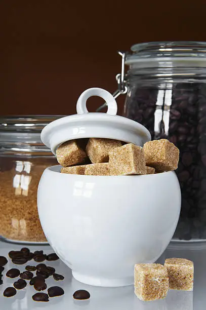 White sugarbowl with brown sugarcubes and jars of coffee and sugar on mixed brown and white background
