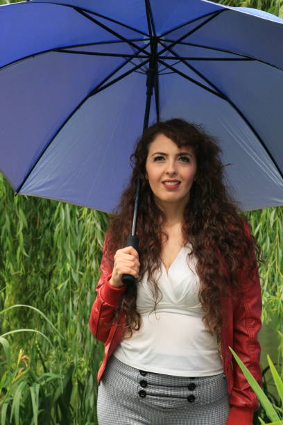 A Mexican model under an umbrella on a rainy day in summer. She is wearing a red leather jacket, white shirt, and gray pants.