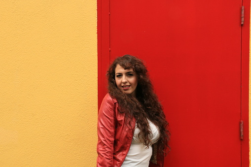 A Mexican model in front of a building with a yellow wall and red door. She is wearing a red leather jacket and white shirt.